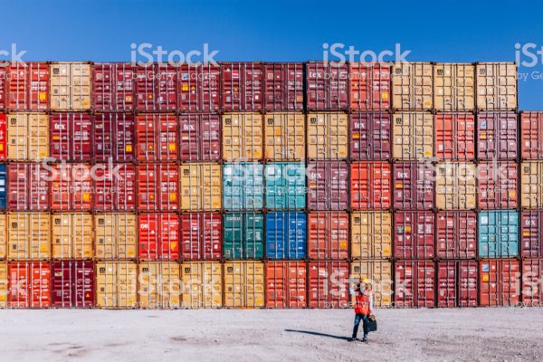 Engineer man with yellow crash helmet and worker west talkig with smartphone and checking cargo freights in front of colorful cargo container stacks in shipping port