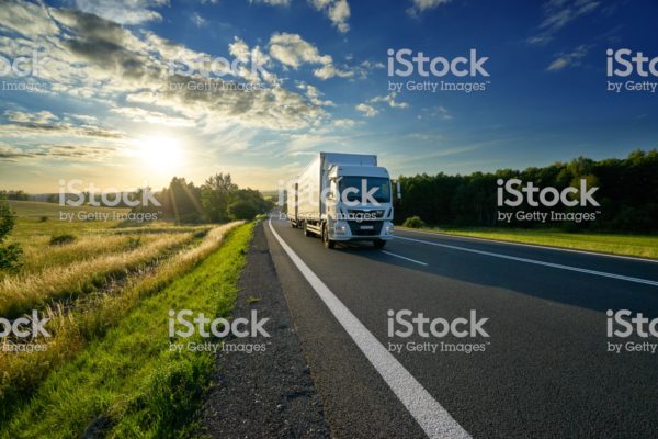 White delivery truck driving on the asphalt road in rural landscape at sunset
